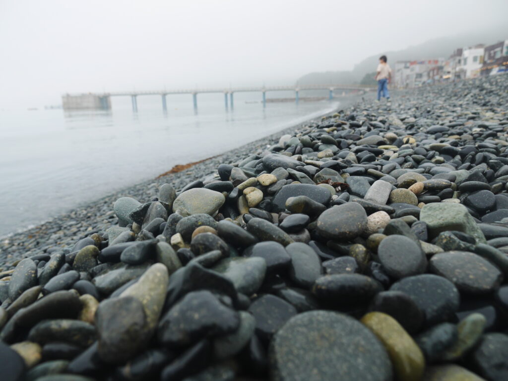 The drizzle hangs in the air but has yet to start falling. The air feels cool but far from unpleasant. The beach lies deserted save us three. There's no sand here, only black and grey pebbles, smooth and round from rolling in the waves. As each wave washes up on the shore it sends a cascade of little stones rattling down the incline towards the water, their noise mixing with wooshing of the ocean; filling the air with a slow rhythm. Being here when no one else is, listening to the crunch of your own footsteps as you're strolling along, picking up a handful of pebbles - they're perfect for throwing - and hurling them into the grey sea just to watch the splash and ripple, feels really meditative.